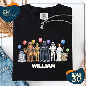A black t-shirt with a graphic design featuring characters from the Star Wars franchise, including Chewbacca, Darth Vader, Stormtrooper, and Yoda, along with colorful balloons.