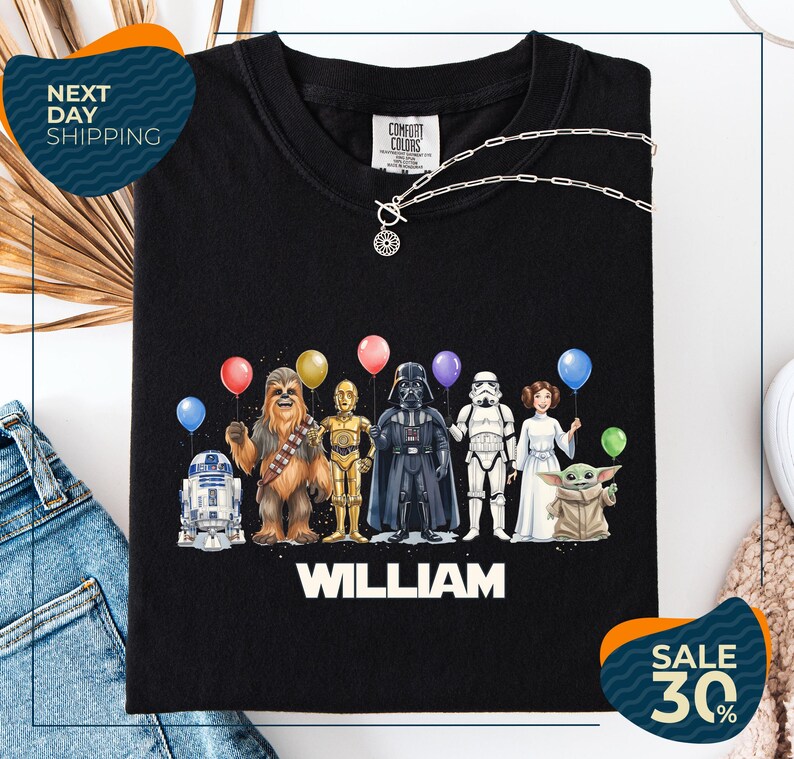 A black t-shirt with a graphic design featuring characters from the Star Wars franchise, including Chewbacca, Darth Vader, Stormtrooper, and Yoda, along with colorful balloons.
