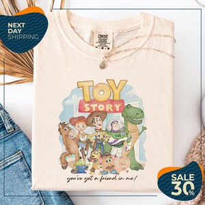 a white t-shirt with a colorful graphic of characters from the Toy Story movie, including Woody, Buzz Lightyear, and other beloved characters. The t-shirt is displayed on a white background, and there are jeans and a sweater visible in the background.