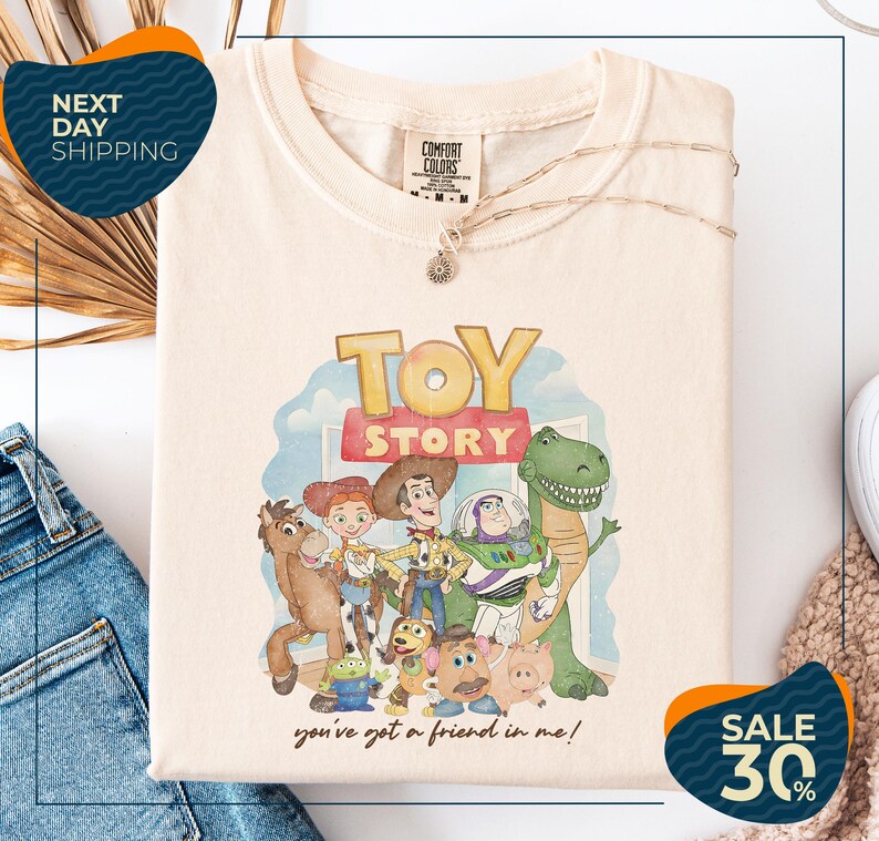 a white t-shirt with a colorful graphic of characters from the Toy Story movie, including Woody, Buzz Lightyear, and other beloved characters. The t-shirt is displayed on a white background, and there are jeans and a sweater visible in the background.