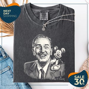a black and white portrait of Walt Disney, with Mickey Mouse on his shoulder, displayed on a gray t-shirt. The t-shirt is folded and placed on a white surface, with a pair of jeans and a beige carpet visible in the background.