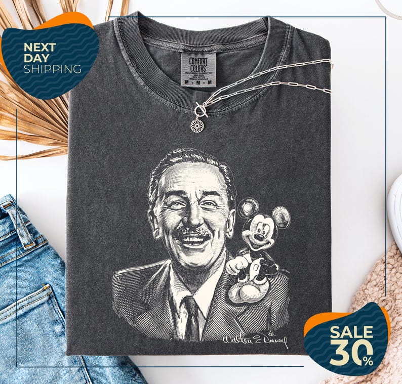 a black and white portrait of Walt Disney, with Mickey Mouse on his shoulder, displayed on a gray t-shirt. The t-shirt is folded and placed on a white surface, with a pair of jeans and a beige carpet visible in the background.