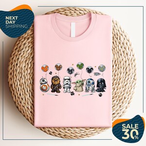 A pink t-shirt with a graphic design featuring various Star Wars characters, including Chewbacca, Stormtrooper, Darth Vader, and Baby Yoda, along with balloons and stars.