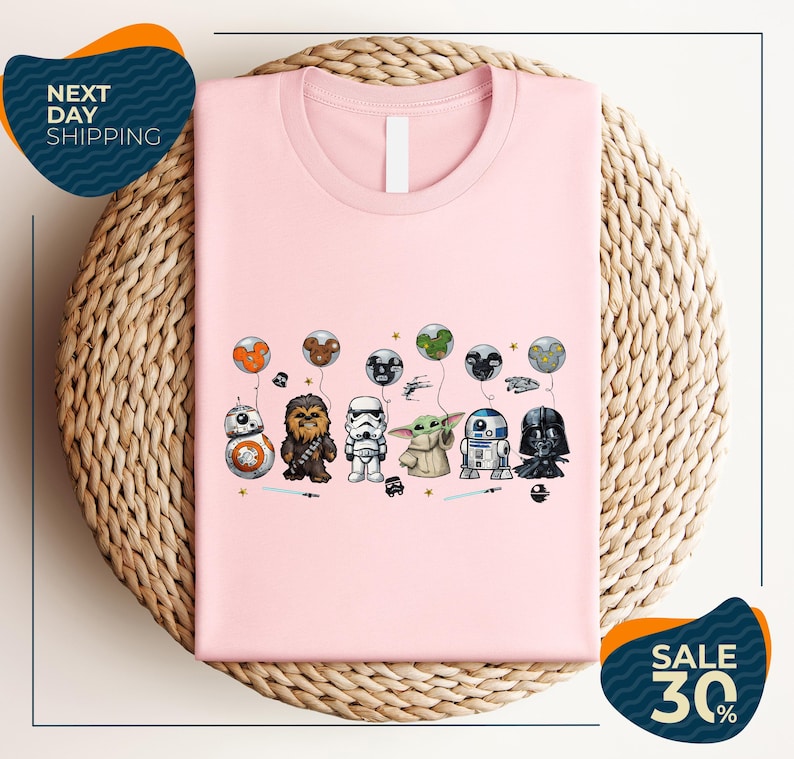 A pink t-shirt with a graphic design featuring various Star Wars characters, including Chewbacca, Stormtrooper, Darth Vader, and Baby Yoda, along with balloons and stars.