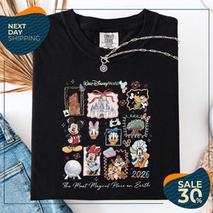 a black t-shirt with a Disney-themed graphic design, including various Disney characters and landmarks. The t-shirt is displayed on a white background, and there are jeans and a necklace visible in the foreground.