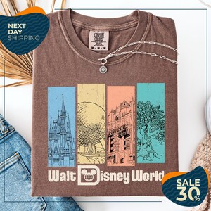 a brown t-shirt with a graphic design of Disney World landmarks, including a castle, roller coaster, and other iconic structures. The t-shirt is displayed on a white background, and there are jeans and a sweater visible in the background.