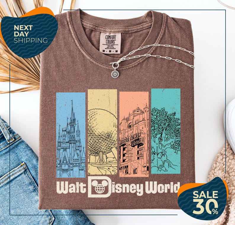 a brown t-shirt with a graphic design of Disney World landmarks, including a castle, roller coaster, and other iconic structures. The t-shirt is displayed on a white background, and there are jeans and a sweater visible in the background.