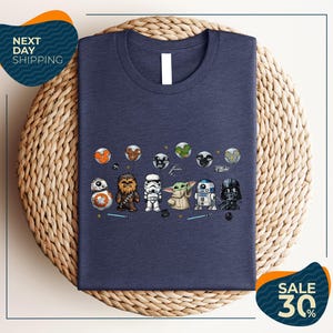 a blue t-shirt with a graphic design of various Star Wars characters, including Chewbacca, Stormtrooper, Baby Yoda, Darth Vader, and others, arranged in a row.