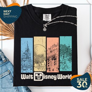 a black t-shirt with a graphic design depicting various landmarks and scenes from Walt Disney World, including a castle, roller coaster, and palm trees. The t-shirt is displayed on a white background, and there are jeans and a necklace visible in the foreground.