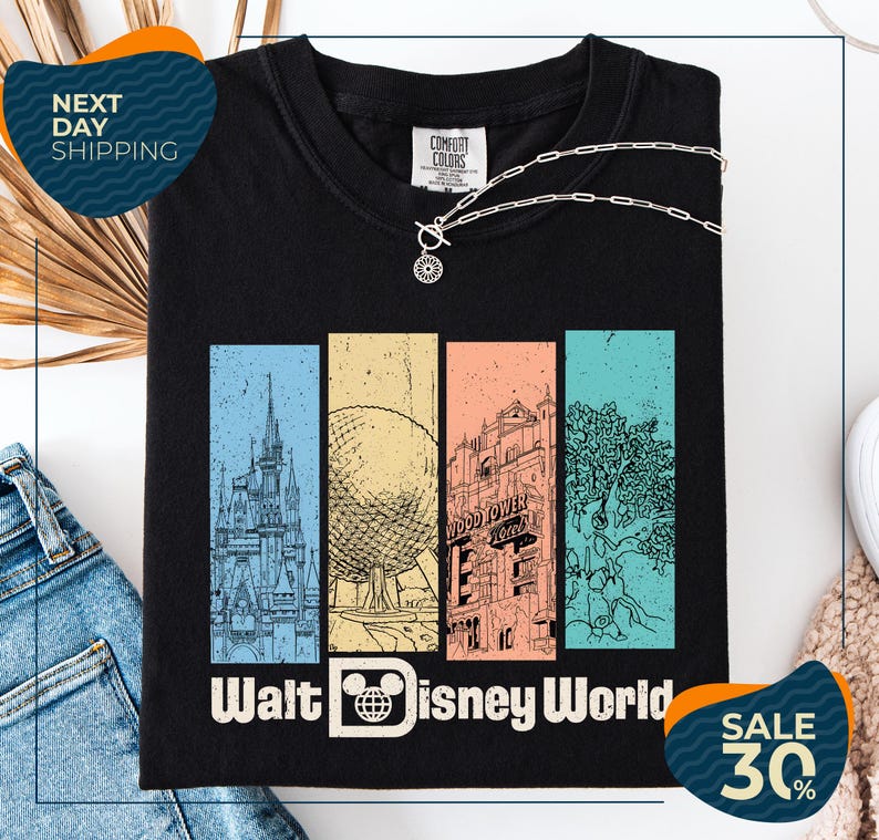 a black t-shirt with a graphic design depicting various landmarks and scenes from Walt Disney World, including a castle, roller coaster, and palm trees. The t-shirt is displayed on a white background, and there are jeans and a necklace visible in the foreground.