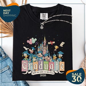 a black t-shirt with a colorful graphic design depicting Disney characters in graduation attire, along with the text "Graduate from the Tassle to the Castle" and the year "2026". The t-shirt is displayed on a white background, and there are jeans and a necklace visible in the foreground.