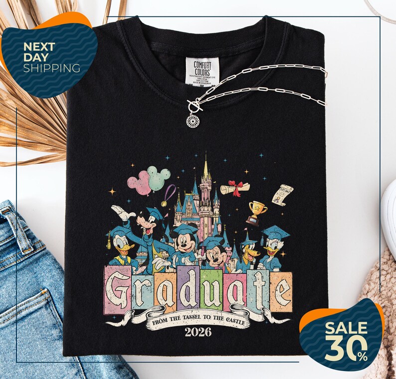 a black t-shirt with a colorful graphic design depicting Disney characters in graduation attire, along with the text "Graduate from the Tassle to the Castle" and the year "2026". The t-shirt is displayed on a white background, and there are jeans and a necklace visible in the foreground.