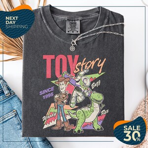 a black t-shirt with a colorful graphic design depicting characters from the Toy Story movie, including Woody and Buzz. The t-shirt is displayed on a white background, and there are jeans and a pair of sunglasses visible in the image.