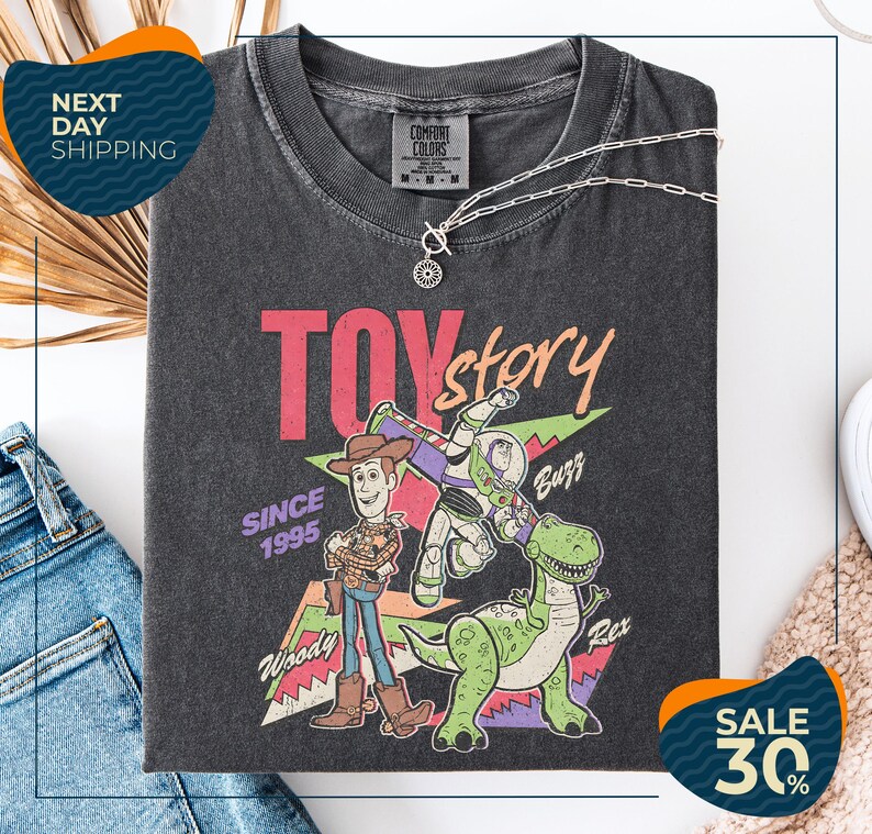 a black t-shirt with a colorful graphic design depicting characters from the Toy Story movie, including Woody and Buzz. The t-shirt is displayed on a white background, and there are jeans and a pair of sunglasses visible in the image.