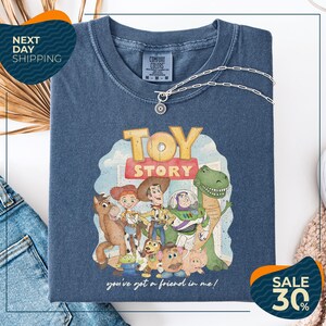a blue t-shirt with a cartoon illustration of characters from the Toy Story movie, including Woody, Buzz Lightyear, and other beloved characters. The t-shirt is displayed on a white background, and there are two sale signs visible, indicating a discount or promotion.
