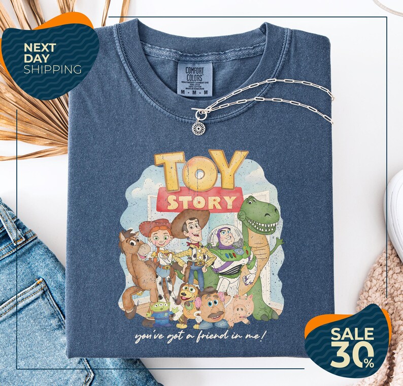 a blue t-shirt with a cartoon illustration of characters from the Toy Story movie, including Woody, Buzz Lightyear, and other beloved characters. The t-shirt is displayed on a white background, and there are two sale signs visible, indicating a discount or promotion.