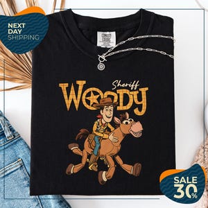 A black t-shirt with a cartoon image of Woody, a character from the Toy Story franchise, and a sheriff riding a horse. The t-shirt is displayed on a white background, and there are jeans and a necklace visible in the image.