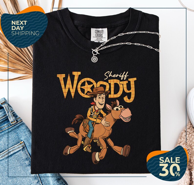 A black t-shirt with a cartoon image of Woody, a character from the Toy Story franchise, and a sheriff riding a horse. The t-shirt is displayed on a white background, and there are jeans and a necklace visible in the image.