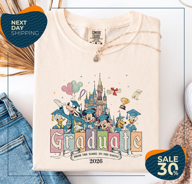 a white t-shirt with a colorful graphic design depicting Disney characters in graduation attire, along with the text "Graduate from the Tasse to the Castle" and the year "2026". The t-shirt is displayed on a white background, and there are jeans visible in the background.