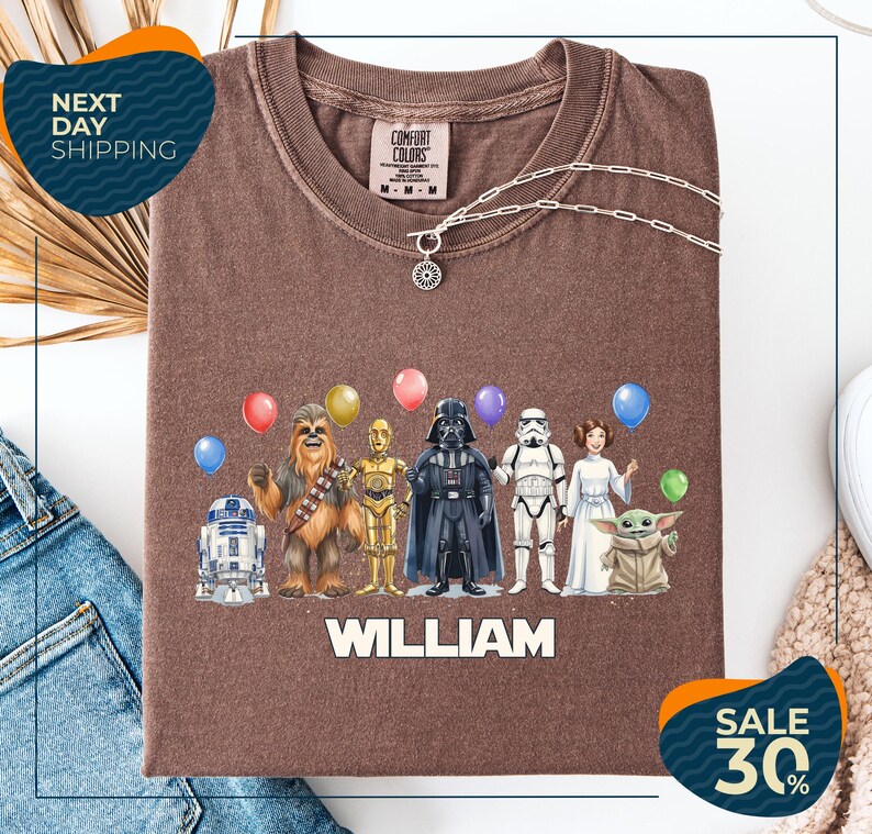 A brown t-shirt with a graphic design featuring various characters from the Star Wars franchise, including Chewbacca, Darth Vader, Stormtrooper, and Yoda, along with colorful balloons. The text "WILLIAM" is displayed below the characters, and there are two sale signs indicating a 30% discount on the t-shirt.