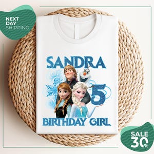 A white t-shirt with a design featuring characters from the Disney movie Frozen, specifically Elsa, Olaf, and Anna, along with the text "SANDRA BIRTHDAY GIRL".