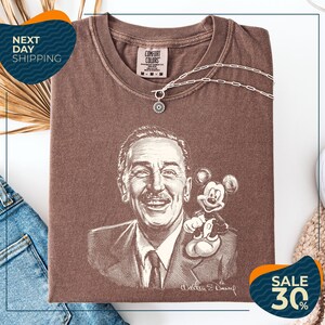 a brown t-shirt with a drawing of a man and a Mickey Mouse, along with a necklace and a chain. The t-shirt is displayed on a white background, and there are some jeans and a sweater visible in the background.