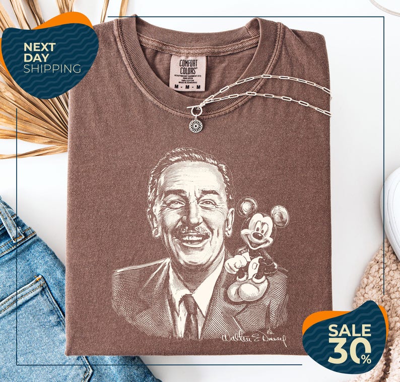 a brown t-shirt with a drawing of a man and a Mickey Mouse, along with a necklace and a chain. The t-shirt is displayed on a white background, and there are some jeans and a sweater visible in the background.