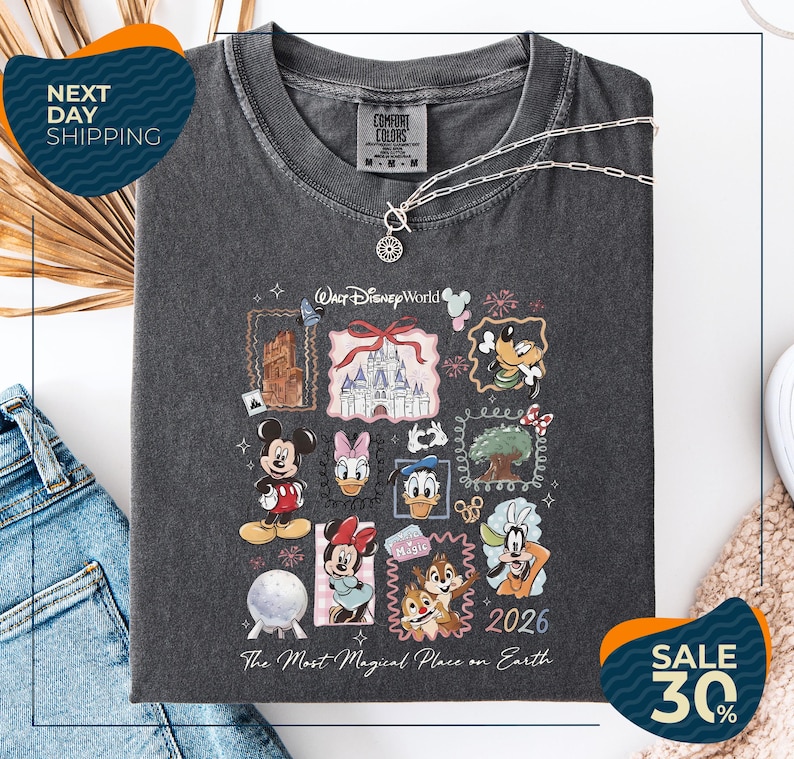 a black t-shirt with a Disney World-themed design, including various Disney characters and landmarks. The t-shirt is displayed on a white background, and there are jeans and a beige blanket visible in the background.