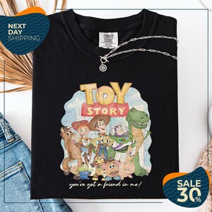 a black t-shirt with a colorful illustration of characters from the Toy Story movie, including Woody, Buzz Lightyear, and other beloved characters. The t-shirt is displayed on a white background, and there are jeans and a necklace visible in the foreground.