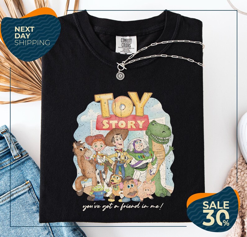 a black t-shirt with a colorful illustration of characters from the Toy Story movie, including Woody, Buzz Lightyear, and other beloved characters. The t-shirt is displayed on a white background, and there are jeans and a necklace visible in the foreground.