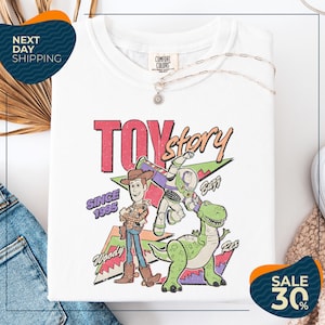 a white t-shirt with a colorful graphic design depicting characters from the Toy Story movie, including Woody and Buzz. The t-shirt is displayed on a white background, and there are some jeans and a necklace visible in the foreground.