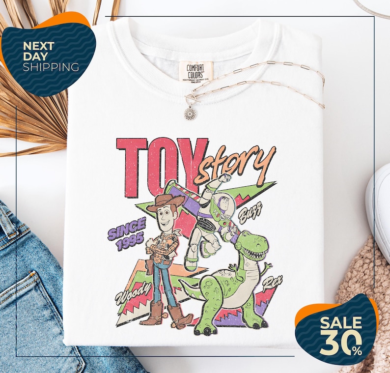 a white t-shirt with a colorful graphic design depicting characters from the Toy Story movie, including Woody and Buzz. The t-shirt is displayed on a white background, and there are some jeans and a necklace visible in the foreground.