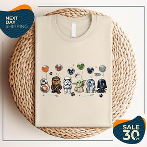 a beige t-shirt with a graphic design of various Star Wars characters, including Chewbacca, Stormtrooper, Darth Vader, and Baby Yoda, along with other Star Wars-themed elements.