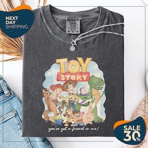 a black t-shirt with a graphic of characters from the Toy Story movie, including Woody, Buzz Lightyear, and other beloved characters. The t-shirt is displayed on a white background, and there are jeans and a necklace visible in the foreground.