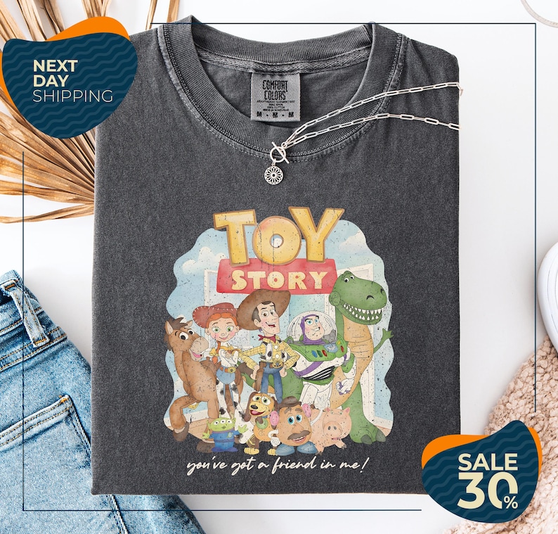 a black t-shirt with a graphic of characters from the Toy Story movie, including Woody, Buzz Lightyear, and other beloved characters. The t-shirt is displayed on a white background, and there are jeans and a necklace visible in the foreground.