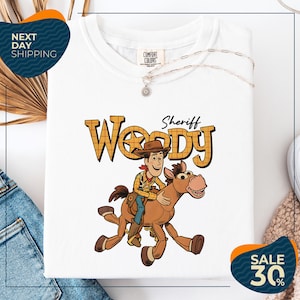 a white t-shirt with a cartoon image of Woody, a character from the Toy Story movie, riding a horse. The t-shirt is displayed on a white background, and there are some jeans and a necklace visible in the background.