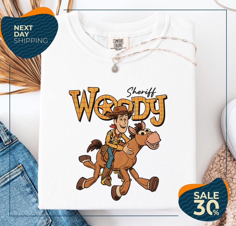 a white t-shirt with a cartoon image of Woody, a character from the Toy Story movie, riding a horse. The t-shirt is displayed on a white background, and there are some jeans and a necklace visible in the background.