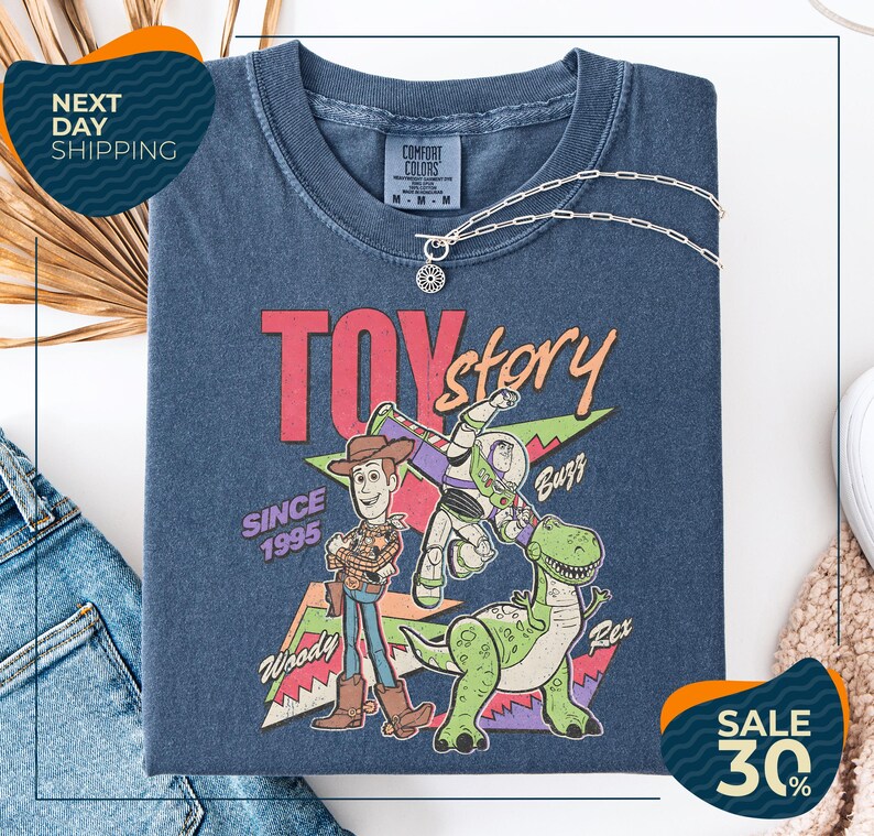 a blue t-shirt with a graphic of Woody and Buzz from Toy Story, along with the text "SINCE 1995" and "Woody & Buzz". The t-shirt is displayed on a white background, and there are jeans and a sweater visible in the background.