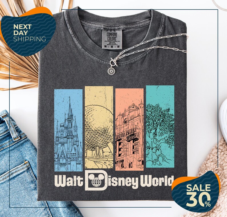 a gray t-shirt with a graphic design of Disney World landmarks, including a castle, roller coaster, and other iconic structures. The t-shirt is displayed on a white background, and there are jeans and a necklace visible in the foreground.