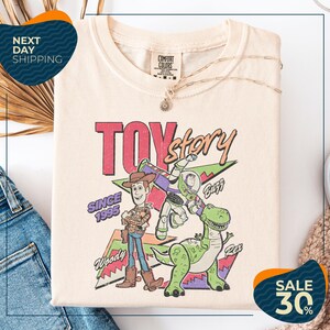 a T-shirt with a colorful graphic of Woody and Buzz from Toy Story, along with the text "Toy Story" and "Since 1995". The T-shirt is displayed on a white background, and there are jeans visible in the background.