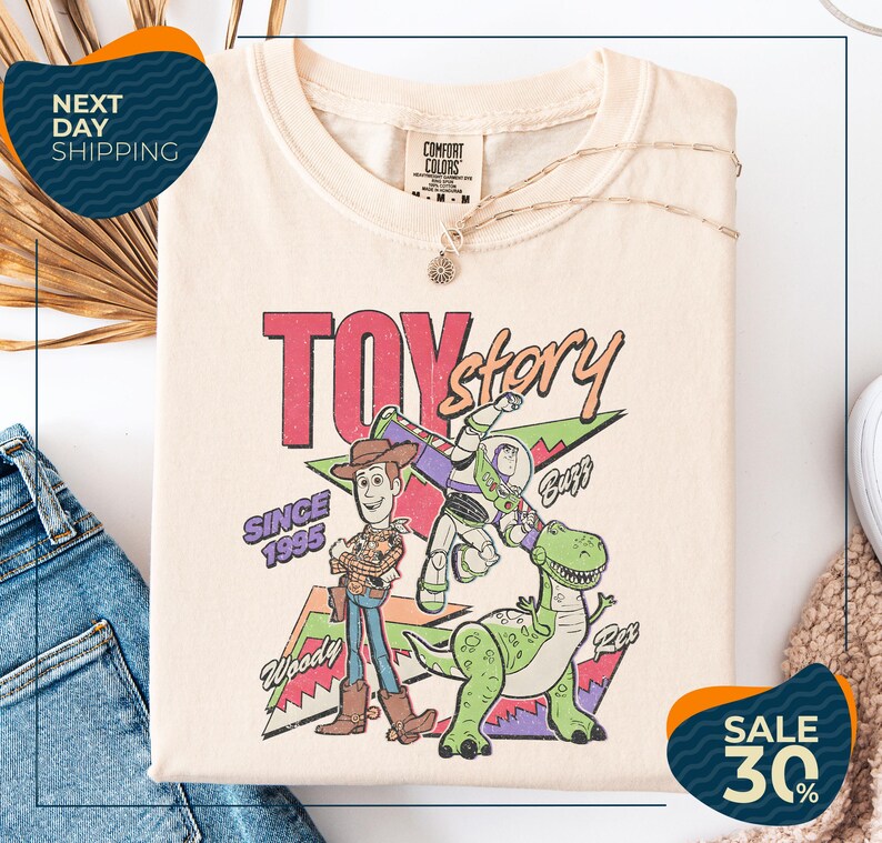 a T-shirt with a colorful graphic of Woody and Buzz from Toy Story, along with the text "Toy Story" and "Since 1995". The T-shirt is displayed on a white background, and there are jeans visible in the background.