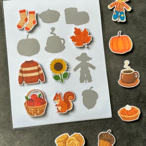 May include: A matching activity sheet featuring a white card with gray silhouettes of autumn items and several colorful die-cut stickers including a scarecrow, pumpkin, coffee mug, pie, acorn, socks, sweater, sunflower, squirrel, apple basket, and hay bales.