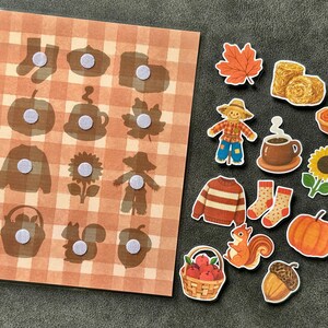 May include: A rectangular board with a light orange and white plaid pattern featuring twelve circular hook-and-loop fasteners, accompanied by twelve separate autumn-themed cutouts including a scarecrow, pumpkin, sunflower, squirrel, acorn, and sweater.