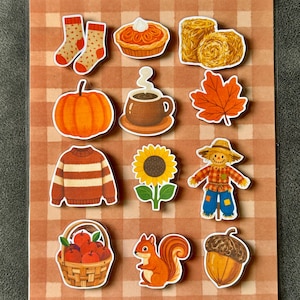 May include: A sticker sheet featuring twelve autumn-themed illustrations including orange socks, a pumpkin pie, hay bales, a pumpkin, a steaming mug, a maple leaf, a striped sweater, a sunflower, a scarecrow, a basket of apples, a squirrel, and an acorn.