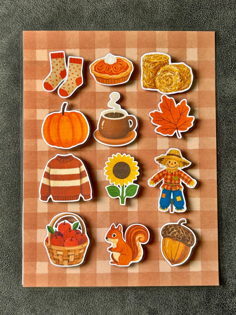 May include: A sticker sheet featuring twelve autumn-themed illustrations including orange socks, a pumpkin pie, hay bales, a pumpkin, a steaming mug, a maple leaf, a striped sweater, a sunflower, a scarecrow, a basket of apples, a squirrel, and an acorn.