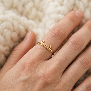 Könnte beinhalten: Goldfarbener Ring mit dem Namen "Amelia" in Schreibschrift. Der Ring befindet sich an einem Finger, vor einem weichen, cremefarbenen Hintergrund.