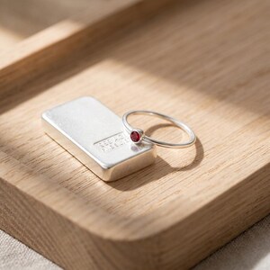 May include: A silver ring with a small, round, red gemstone sits next to a silver bar on a wooden surface. The ring has a thin band and a simple design. The silver bar has text and markings on its surface.