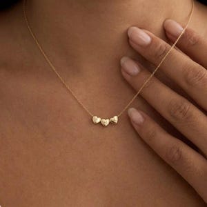 May include: A delicate gold necklace featuring three heart-shaped pendants. Each heart pendant has a single initial engraved on it. The necklace is displayed on a person's neck and chest.