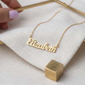 May include: A gold-colored necklace with the name "Elizabeth" in a cursive font. The necklace is displayed on a white cloth, with a small gold cube in the foreground. The chain is delicate and the pendant is shiny.