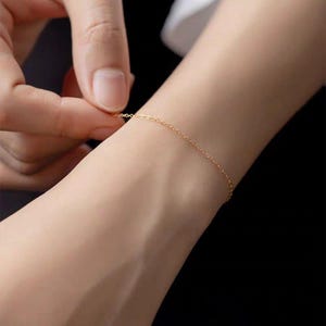 May include: A delicate gold chain bracelet. The fine chain is draped across a wrist, with a hand adjusting the clasp. The bracelet is a simple, elegant piece of jewelry.